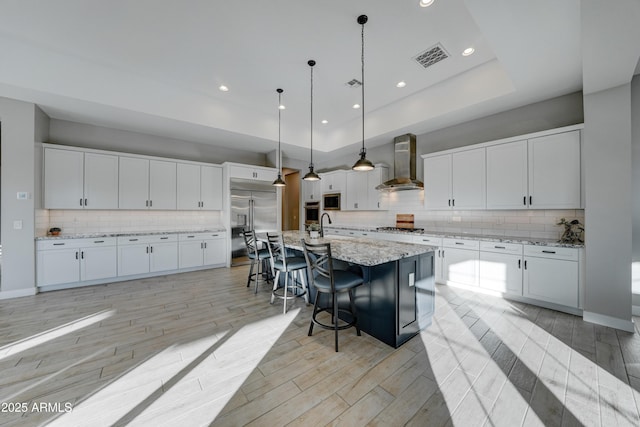 kitchen with pendant lighting, wall chimney range hood, appliances with stainless steel finishes, a kitchen island with sink, and white cabinetry