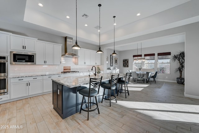 kitchen featuring white cabinetry, decorative light fixtures, stainless steel appliances, a kitchen island with sink, and wall chimney range hood