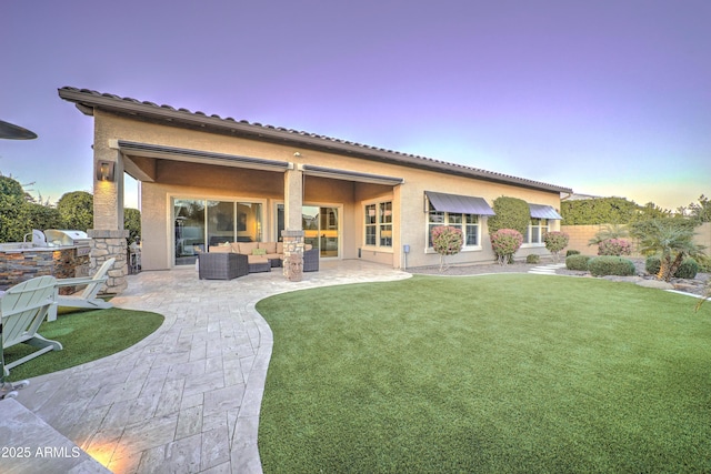 back house at dusk featuring a patio, an outdoor kitchen, an outdoor hangout area, and a lawn