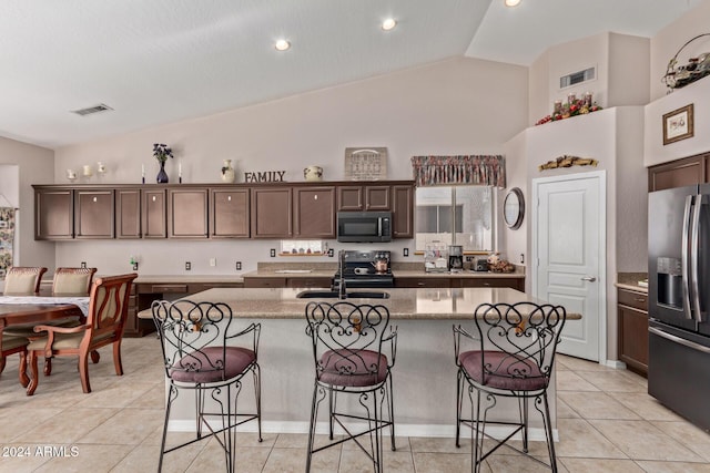 kitchen with appliances with stainless steel finishes, dark brown cabinetry, vaulted ceiling, a kitchen island with sink, and light tile patterned floors