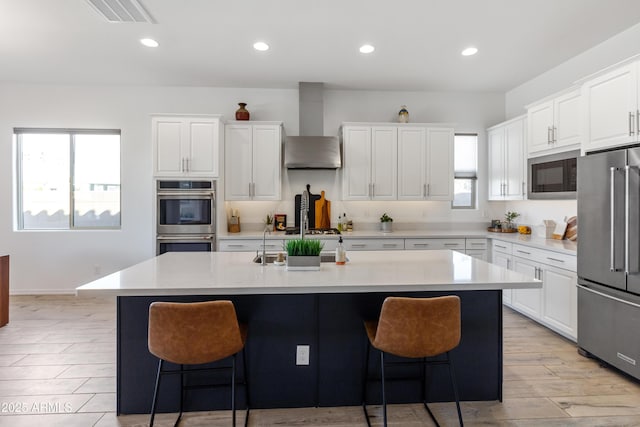 kitchen with stainless steel appliances, an island with sink, white cabinets, and wall chimney exhaust hood