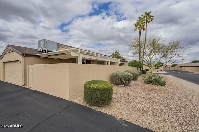 view of side of home featuring central AC unit, fence, stucco siding, a garage, and aphalt driveway