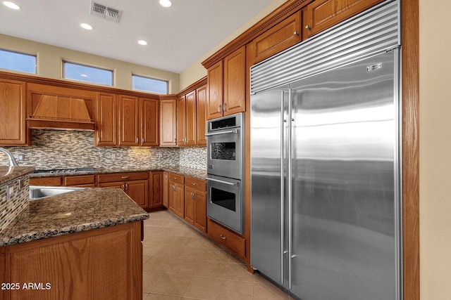 kitchen featuring appliances with stainless steel finishes, dark stone countertops, tasteful backsplash, light tile patterned flooring, and custom exhaust hood