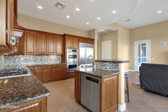 kitchen featuring a breakfast bar area, dark stone countertops, backsplash, stainless steel appliances, and an island with sink