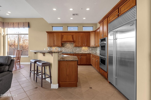 kitchen with appliances with stainless steel finishes, dark stone countertops, a kitchen breakfast bar, decorative backsplash, and custom range hood