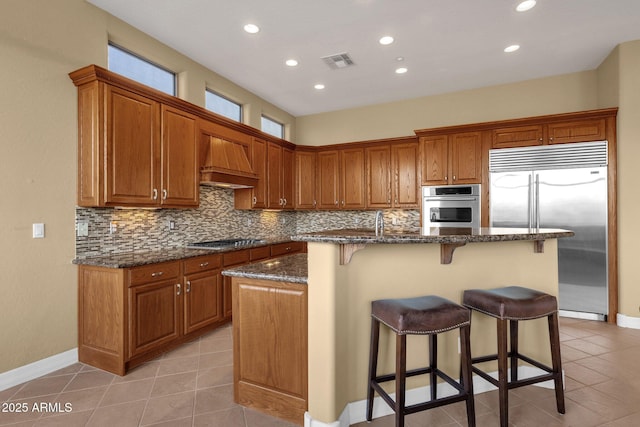 kitchen featuring wall chimney exhaust hood, appliances with stainless steel finishes, an island with sink, dark stone counters, and backsplash