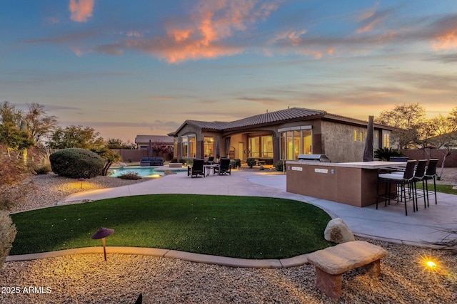 back house at dusk featuring a patio, exterior bar, a yard, and an outdoor kitchen