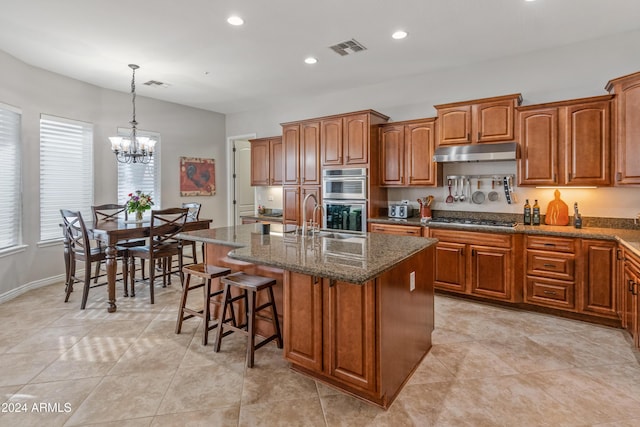 kitchen featuring stainless steel appliances, decorative light fixtures, dark stone countertops, a chandelier, and an island with sink