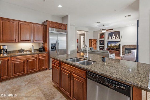 kitchen featuring dark stone counters, stainless steel appliances, ceiling fan, sink, and a center island with sink