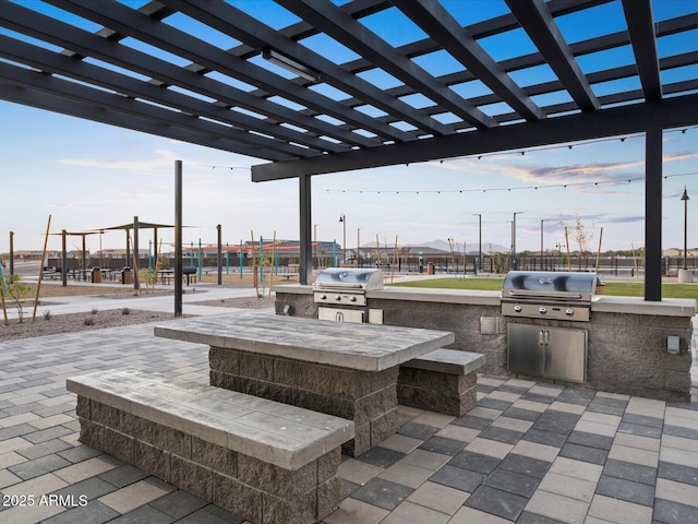 patio terrace at dusk featuring exterior kitchen, area for grilling, and a pergola
