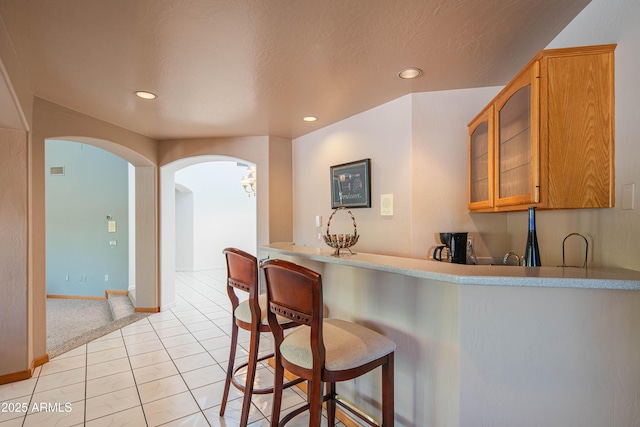 kitchen with a breakfast bar, arched walkways, brown cabinetry, light tile patterned floors, and glass insert cabinets
