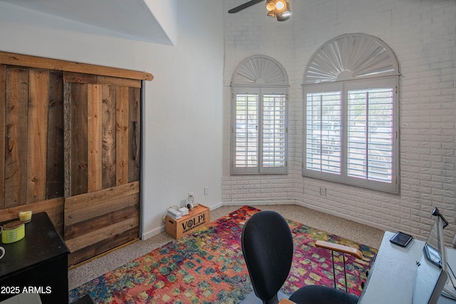 carpeted office featuring baseboards, a ceiling fan, and brick wall