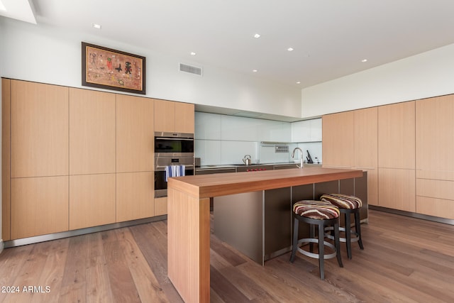 kitchen featuring double oven, light brown cabinets, an island with sink, and light wood-type flooring