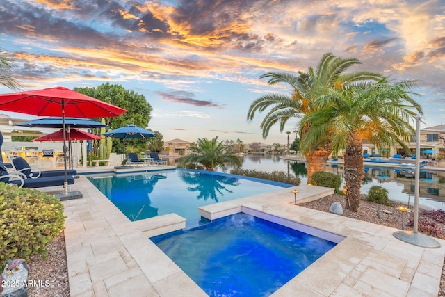 pool at dusk with an in ground hot tub, a water view, and a patio