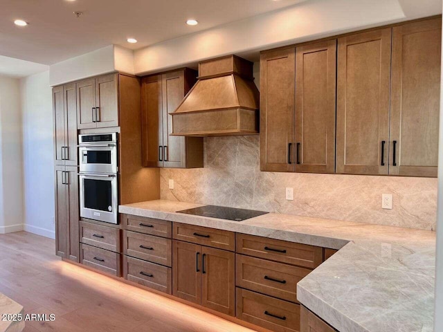 kitchen with black electric stovetop, premium range hood, double oven, light countertops, and decorative backsplash