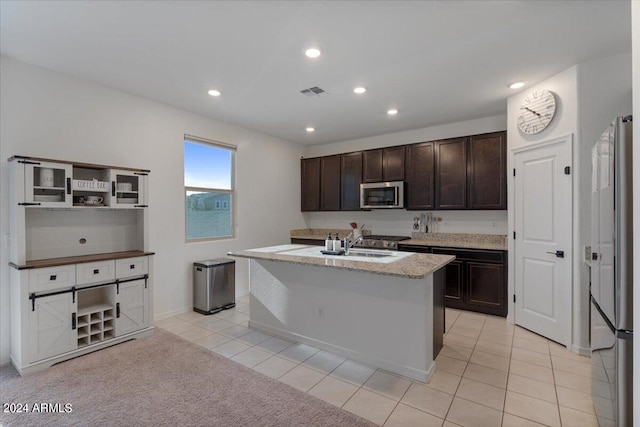 kitchen with light tile patterned floors, stainless steel appliances, an island with sink, sink, and dark brown cabinetry