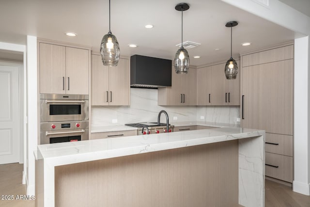 kitchen with light brown cabinets, wall chimney range hood, decorative backsplash, light stone countertops, and decorative light fixtures