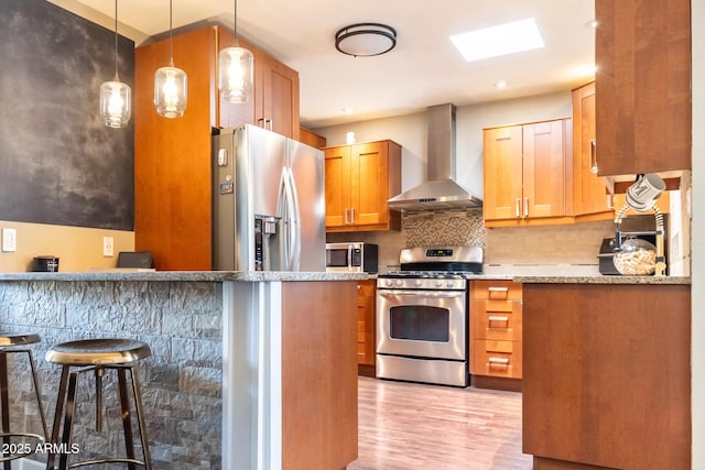 kitchen featuring tasteful backsplash, wall chimney range hood, light wood-type flooring, appliances with stainless steel finishes, and a kitchen bar
