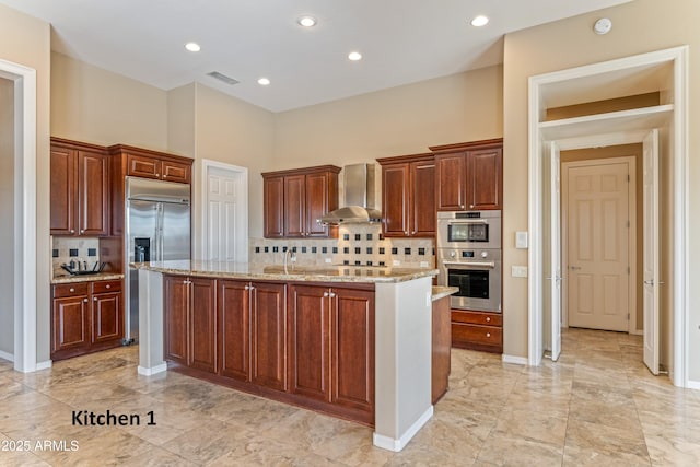 kitchen with light stone countertops, appliances with stainless steel finishes, wall chimney range hood, tasteful backsplash, and an island with sink