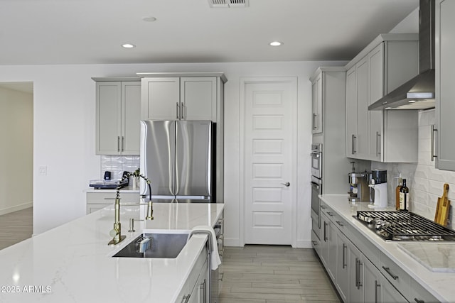 kitchen with appliances with stainless steel finishes, light stone counters, gray cabinets, wall chimney range hood, and a sink