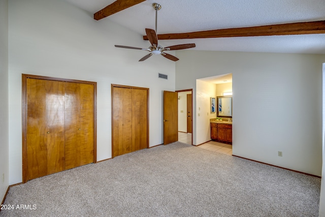 unfurnished bedroom with beam ceiling, ensuite bathroom, multiple closets, and light colored carpet