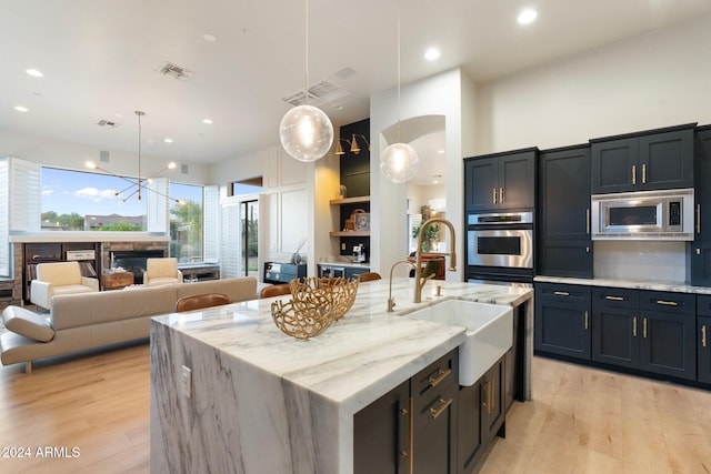 kitchen with light stone countertops, hanging light fixtures, a center island with sink, appliances with stainless steel finishes, and light wood-type flooring