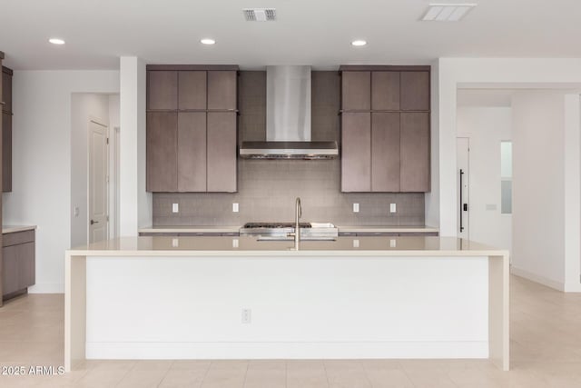 kitchen with a kitchen island with sink, light countertops, wall chimney range hood, and decorative backsplash