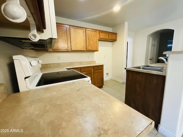 kitchen with a sink, arched walkways, brown cabinetry, light tile patterned floors, and white electric range
