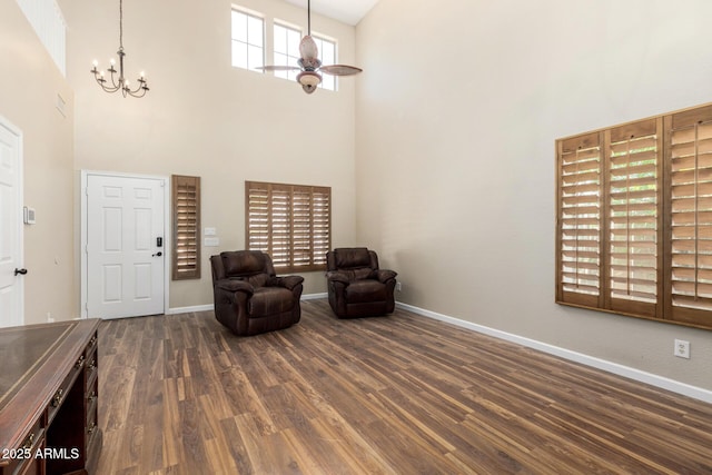 living area featuring baseboards, ceiling fan with notable chandelier, dark wood-style floors, and a towering ceiling