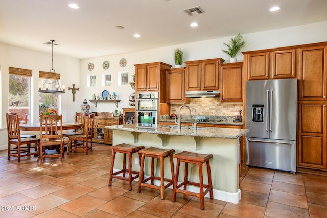 kitchen with tasteful backsplash, a center island with sink, pendant lighting, stainless steel appliances, and light stone countertops