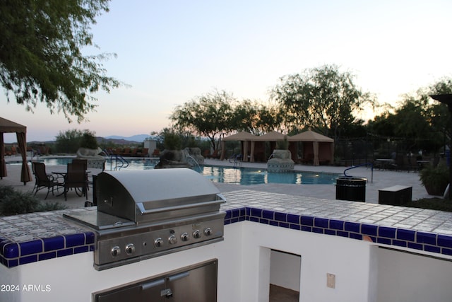 patio terrace at dusk with exterior kitchen, a community pool, and grilling area