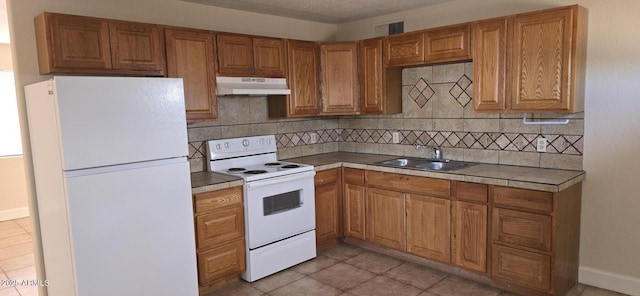 kitchen with under cabinet range hood, white appliances, a sink, tasteful backsplash, and brown cabinetry