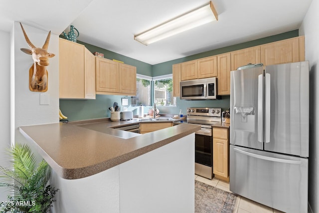 kitchen featuring light tile patterned floors, appliances with stainless steel finishes, a peninsula, light brown cabinetry, and a sink