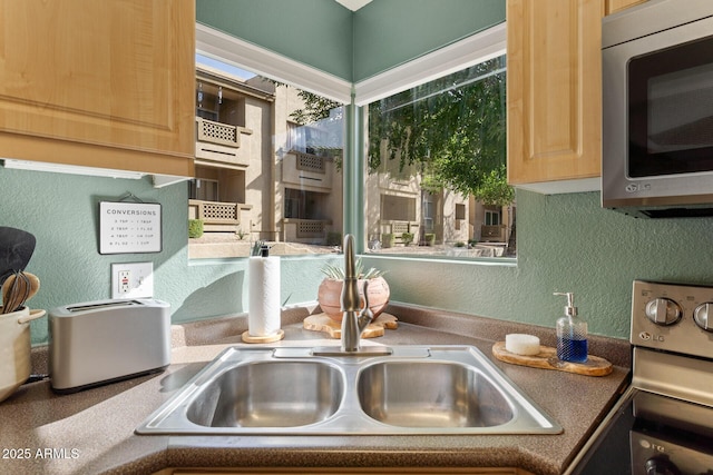 kitchen featuring a textured wall, a sink, range, light brown cabinetry, and stainless steel microwave