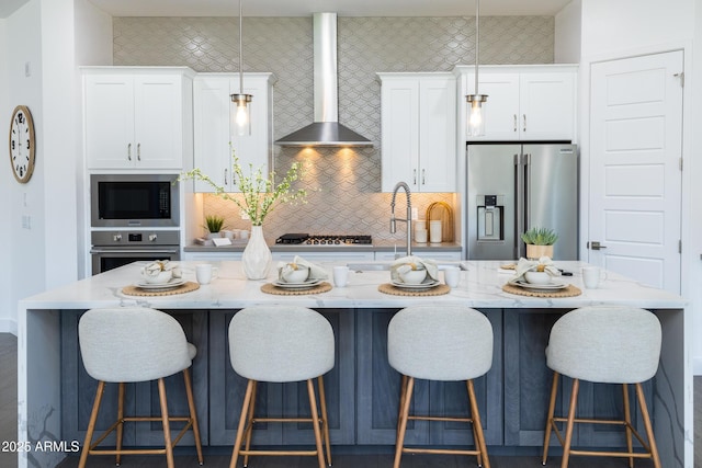 kitchen featuring wall chimney exhaust hood, a kitchen island with sink, white cabinetry, stainless steel appliances, and light stone counters