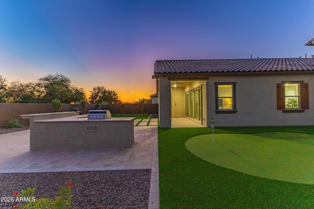 patio terrace at dusk with a fenced backyard, a grill, and an outdoor kitchen