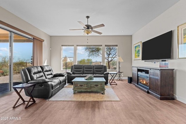 living area with a glass covered fireplace, baseboards, light wood-type flooring, and a ceiling fan