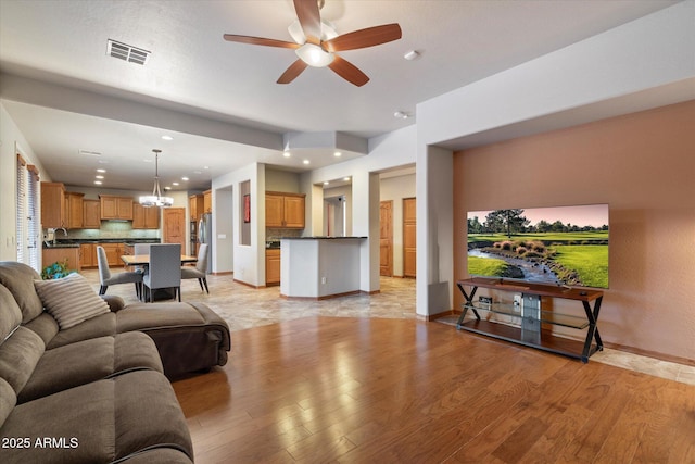 living area featuring light wood-type flooring, baseboards, visible vents, and ceiling fan with notable chandelier