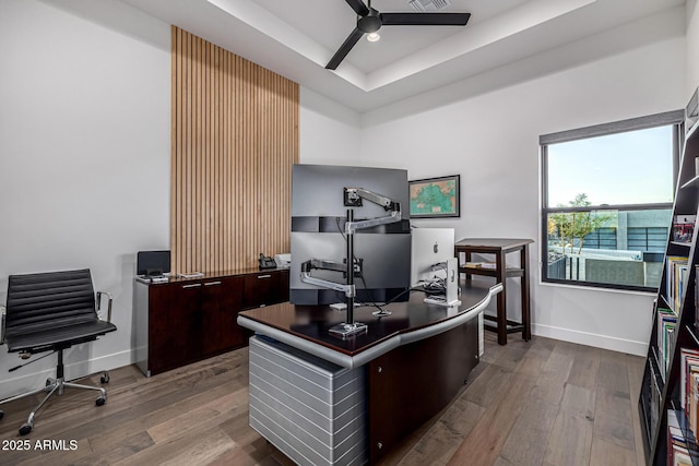 office space featuring a ceiling fan, a tray ceiling, baseboards, and wood finished floors