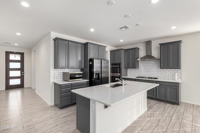 kitchen with tasteful backsplash, wall chimney exhaust hood, stainless steel appliances, and an island with sink