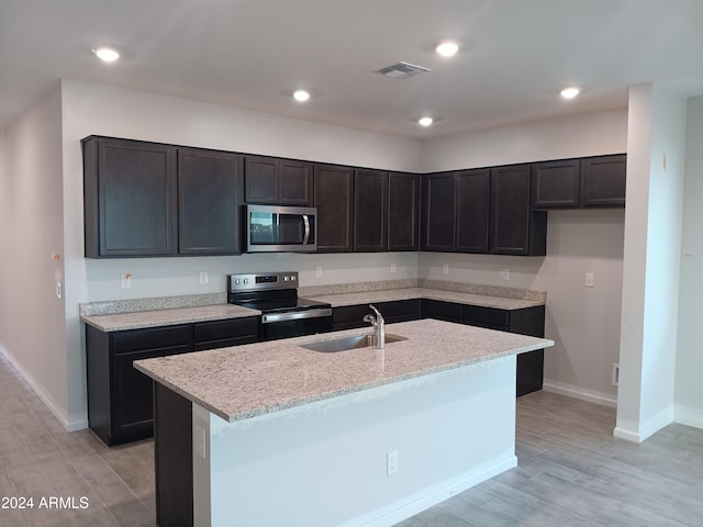 kitchen with appliances with stainless steel finishes, sink, light stone counters, light hardwood / wood-style flooring, and a center island with sink