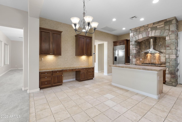 kitchen with wall chimney range hood, black gas cooktop, stainless steel built in refrigerator, dark brown cabinetry, and built in desk