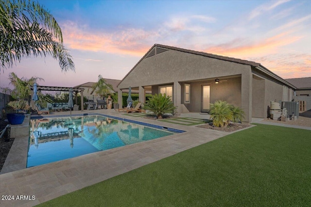 pool at dusk with a pergola, a patio area, ceiling fan, and a lawn