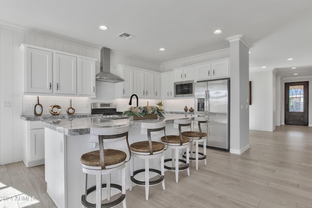 kitchen with stainless steel appliances, wall chimney range hood, an island with sink, a breakfast bar, and white cabinets