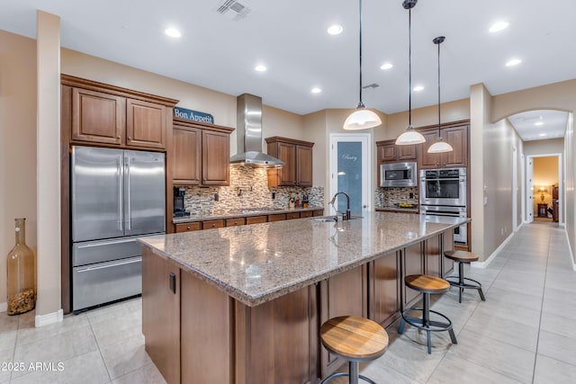 kitchen with wall chimney exhaust hood, tasteful backsplash, a kitchen island with sink, stone counters, and stainless steel appliances