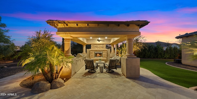 patio terrace at dusk featuring an outdoor stone fireplace, a lawn, and ceiling fan