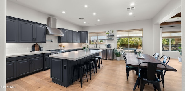 kitchen with wall chimney range hood, backsplash, a kitchen bar, light hardwood / wood-style floors, and a center island