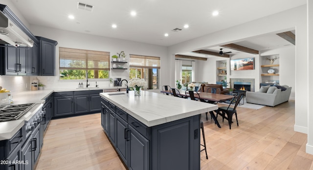 kitchen with wall chimney range hood, beam ceiling, light hardwood / wood-style flooring, a center island with sink, and sink