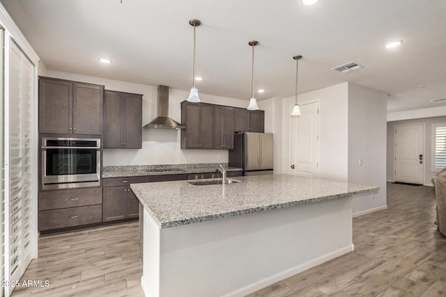 kitchen featuring wall chimney range hood, sink, light stone countertops, an island with sink, and appliances with stainless steel finishes