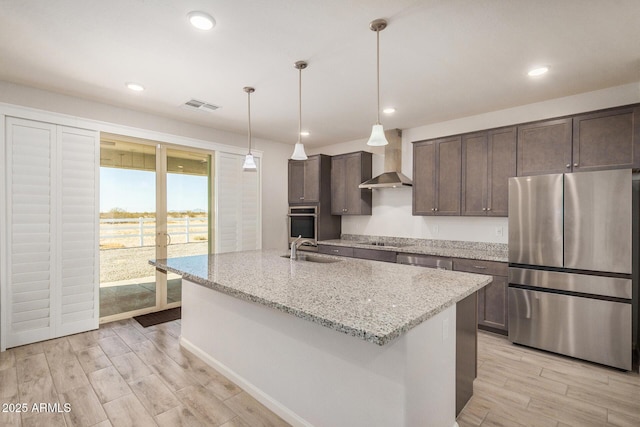 kitchen featuring stainless steel appliances, pendant lighting, wall chimney exhaust hood, light stone counters, and sink